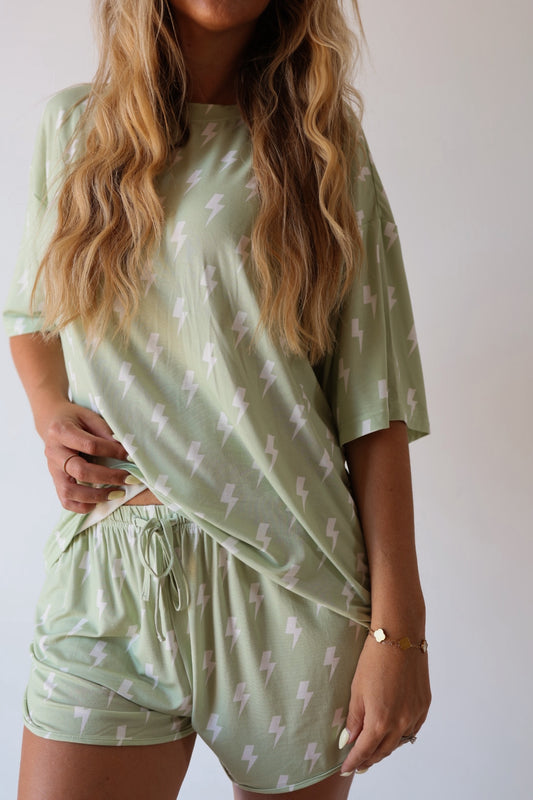 A woman with long wavy hair wears the forever french baby Women's Bamboo Short Set | Flash, a breathable, hypo-allergenic light green pajama set with white lightning bolts. The loose short-sleeve top and drawstring shorts offer comfy loungewear.