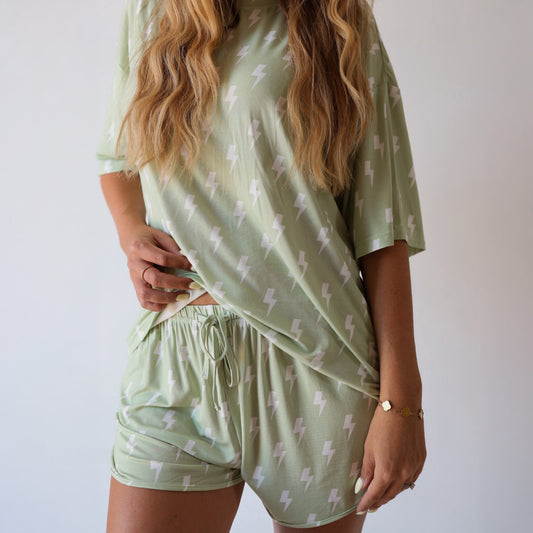 A woman wears the forever french baby Women's Bamboo Short Set | Flash—a light green, bamboo pajama set with a white lightning bolt print, featuring a loose short-sleeve top and drawstring shorts—standing against a plain white background.