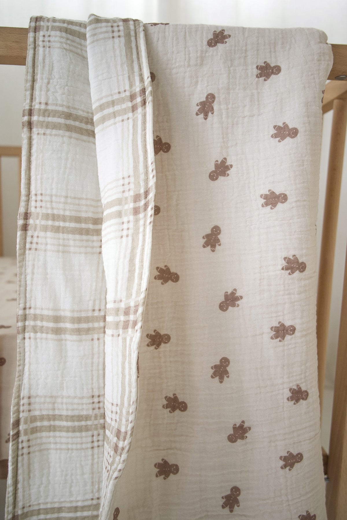 A reversible Muslin Quilt | Gingersnap & Ellis Plaid draped over a crib rail, featuring one side with a brown gingerbread man print and the other with a neutral plaid pattern.