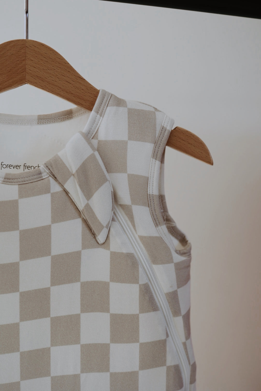 The forever french baby Bamboo Sleep Sack | Checkerboard, in beige and white checks, hangs on a wooden hanger and features a zipper with a snap button on the shoulder.