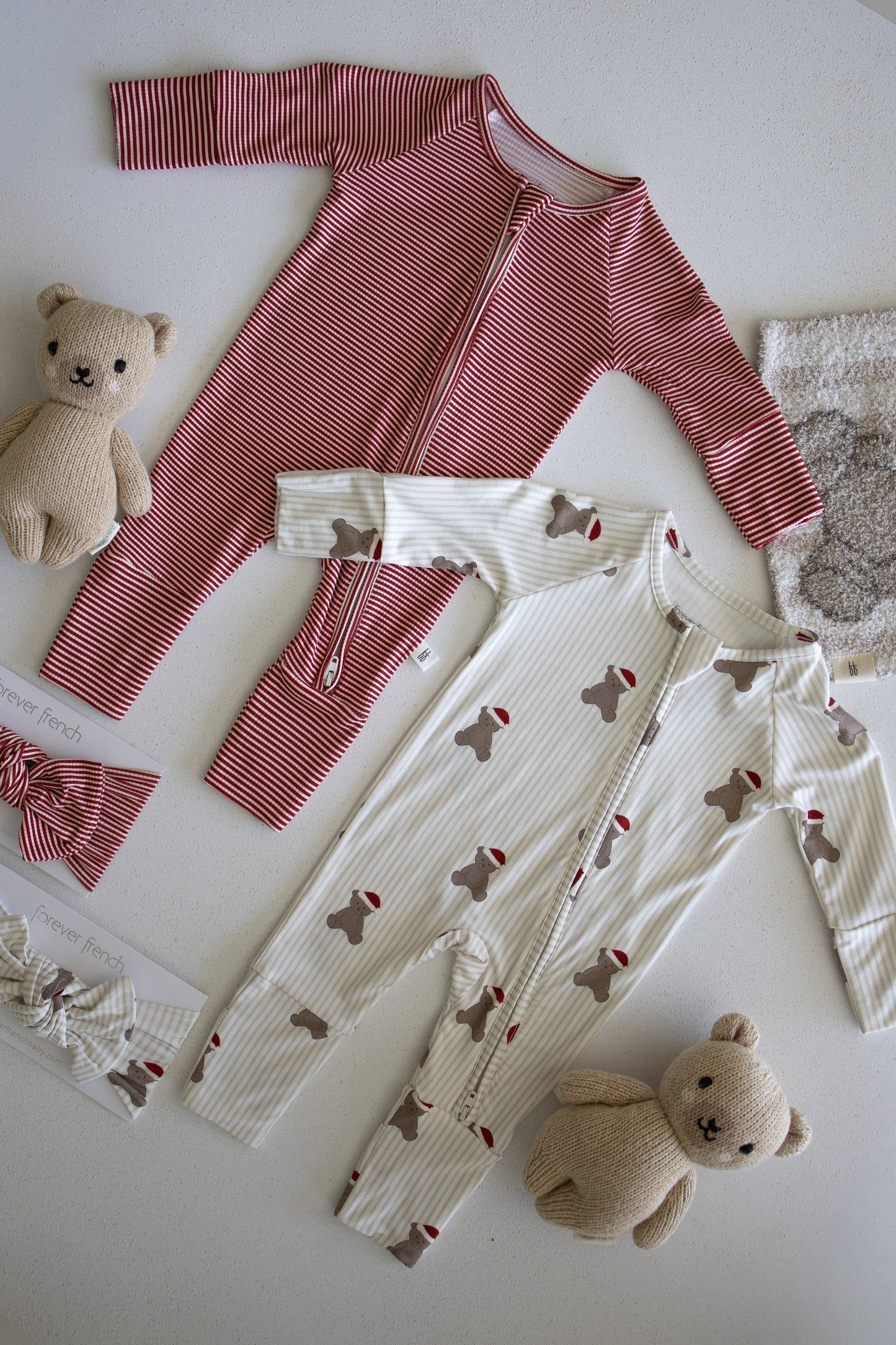 Two Bamboo Zip Pajamas from forever french baby—one in red stripes and the other featuring a whimsical Santa Teddy print—are displayed on a light surface, accompanied by two plush teddy bears, a coordinating headband, and a piece of textured fabric. These hypoallergenic pajamas provide both cozy comfort and charming style for your little one.