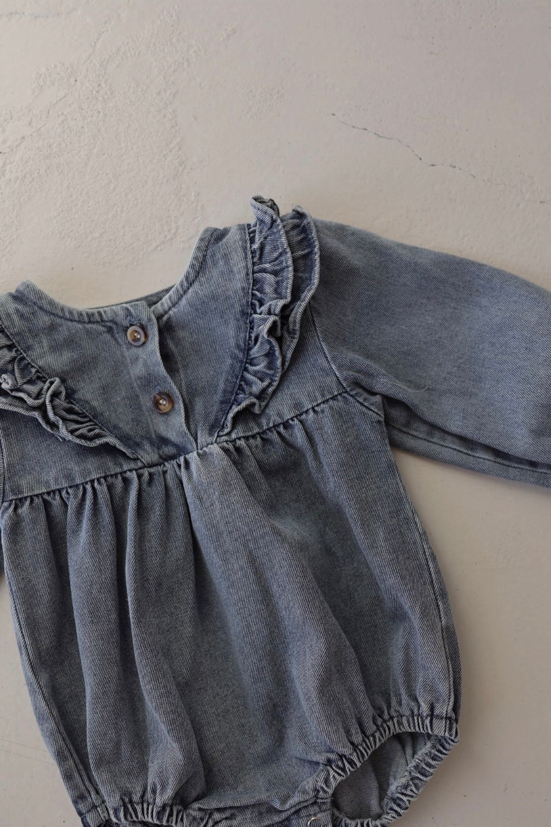 The Denim Romper | Ruffle by forever french baby is a blue, long-sleeved tunic-style romper with chest ruffles and front buttons, shown laid flat on a light textured surface.