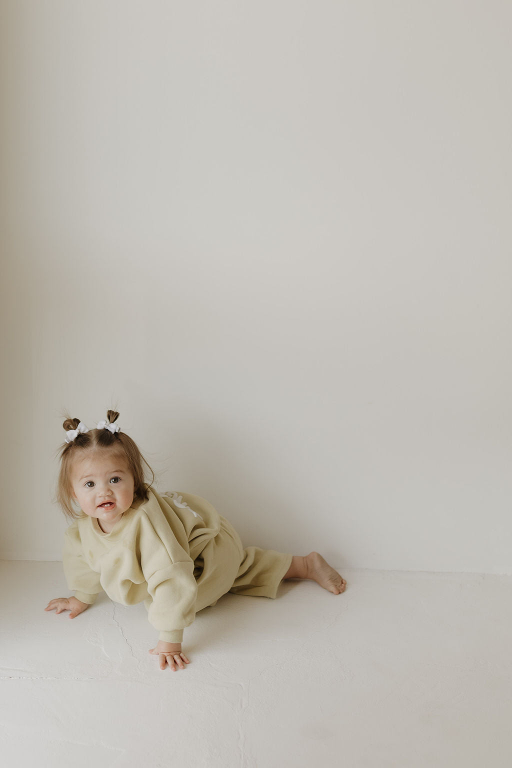 A young child with two small hair buns crawls on a light-colored floor, dressed in the "Child Sweat Set | All the Good Things" by forever french baby. The background is plain and softly illuminated, creating a serene atmosphere typical of Forever French Baby's timeless elegance.