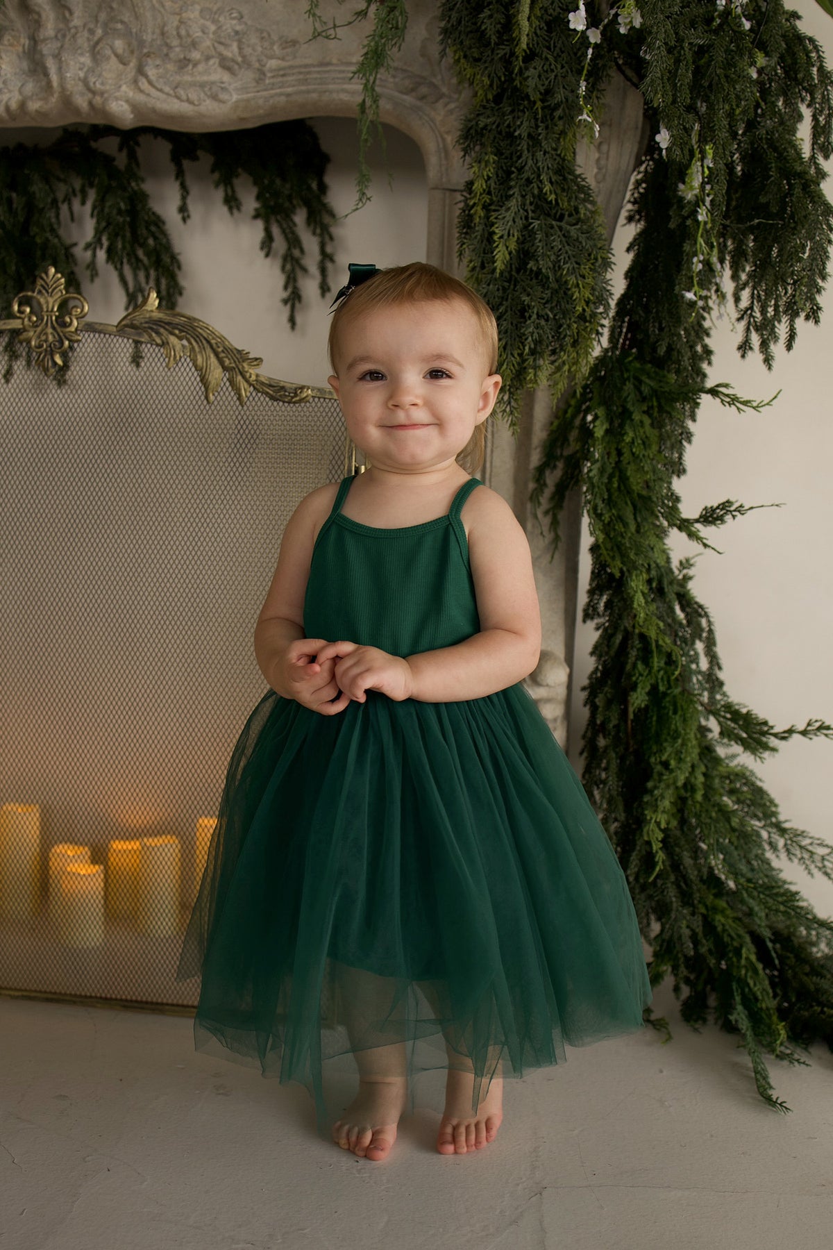 A toddler stands in front of a fireplace wearing a dark green ff Tutu Dress | Mistletoe with a layered tulle skirt.