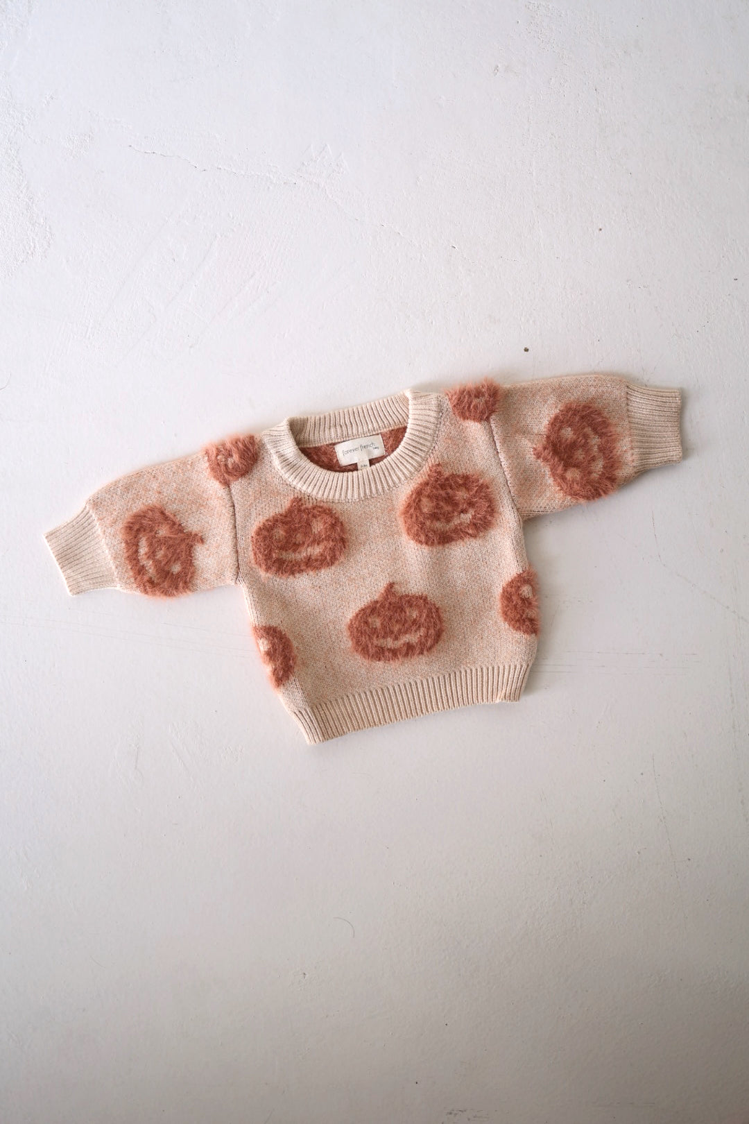 A small, beige knitted sweater with long sleeves from forever french baby, adorned with large, fluffy, rust-colored flower or cloud-like patterns. This Cozy Sweater | Pumpkin is laid flat on a white textured surface—an autumn essential for any wardrobe.