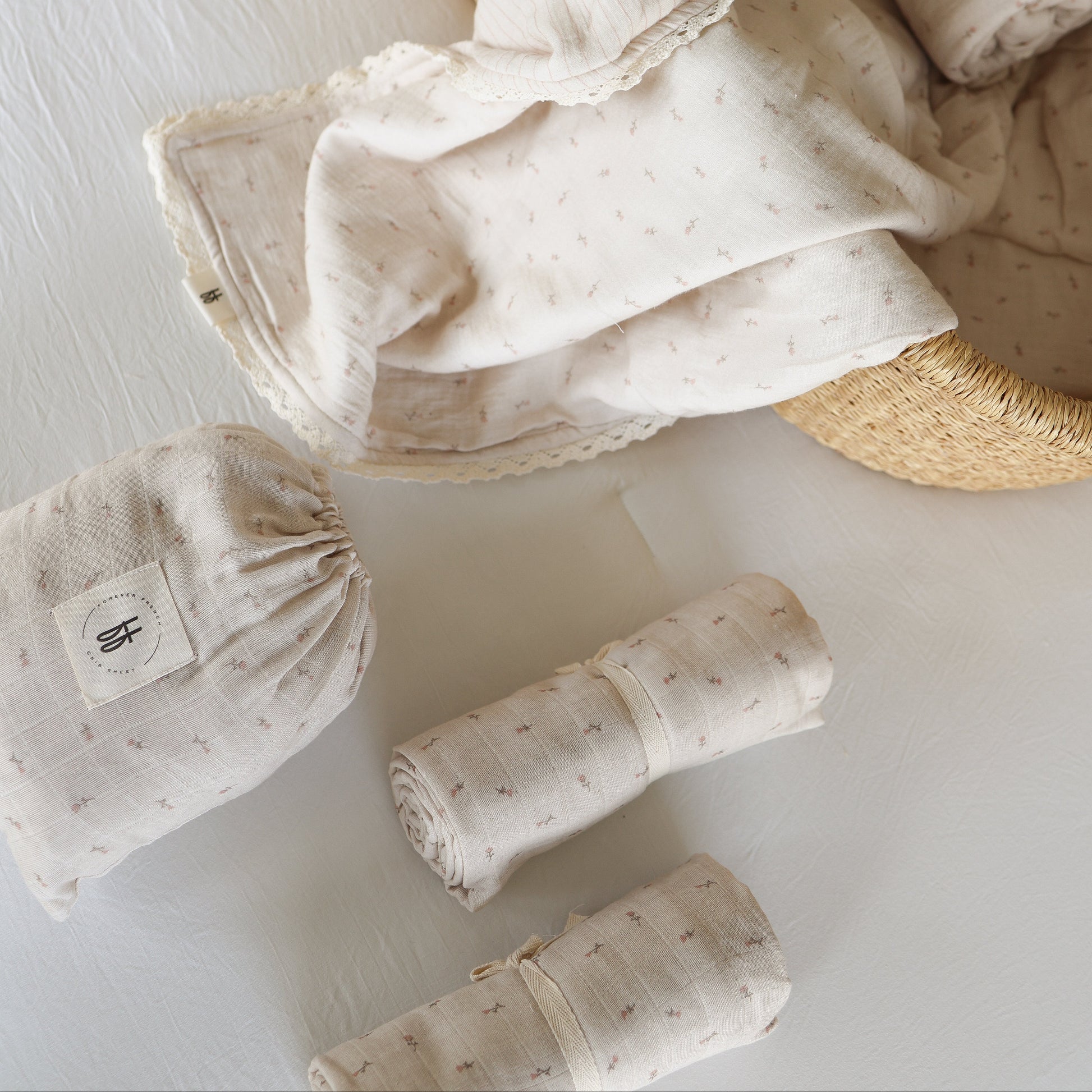 The Muslin Swaddle | Lucy by forever french baby, featuring a soft cream hue with a subtle star pattern, is draped over a woven basket, with two rolled swaddles and a matching drawstring pouch nearby.