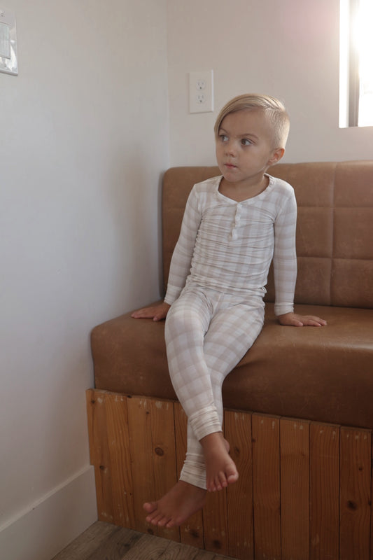 A young blonde child in forever french baby’s Bamboo Two Piece Pajamas | Gingham sits barefoot on a brown cushioned bench against a light wall, gazing thoughtfully to the side.