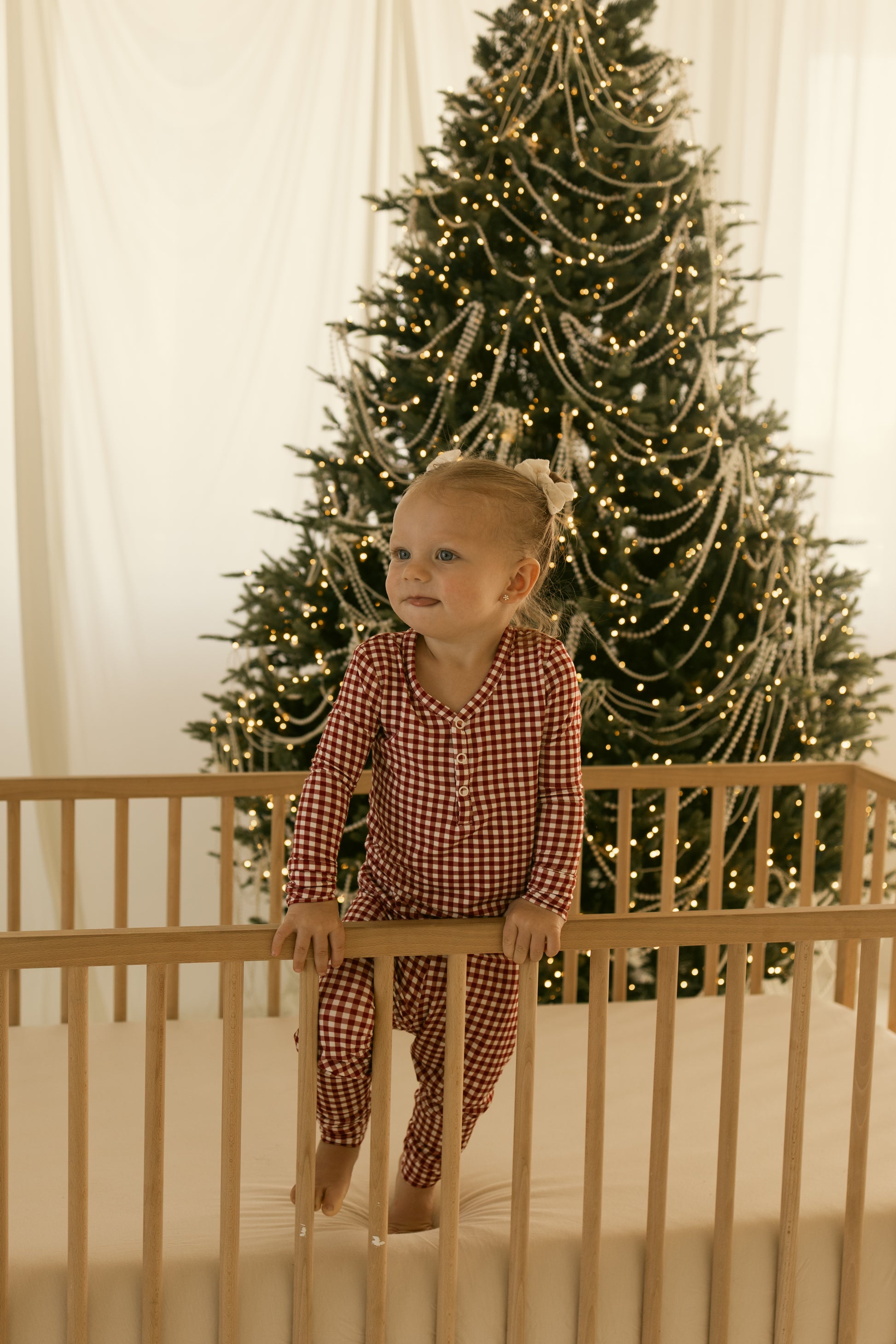 A woman wearing forever french baby’s Women’s Bamboo Pajamas in Red Gingham stands by a wooden crib, with a bright Christmas tree decorated with lights and garlands behind her, as soft natural light fills the festive room.