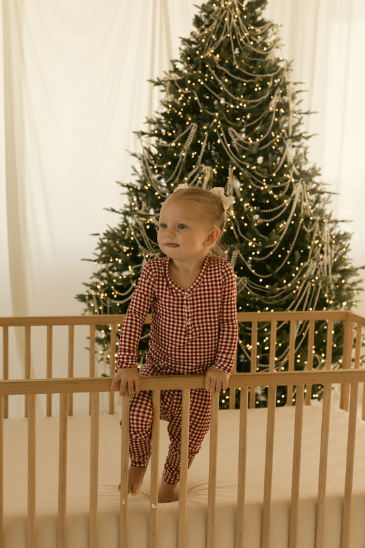 A woman wearing forever french baby’s Women’s Bamboo Pajamas in Red Gingham stands by a wooden crib, with a bright Christmas tree decorated with lights and garlands behind her, as soft natural light fills the festive room.