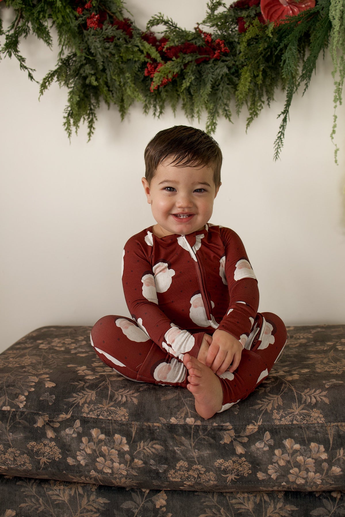 A smiling toddler sits on a patterned ottoman wearing forever french baby Bamboo Zip Pajamas | Vintage Santa.
