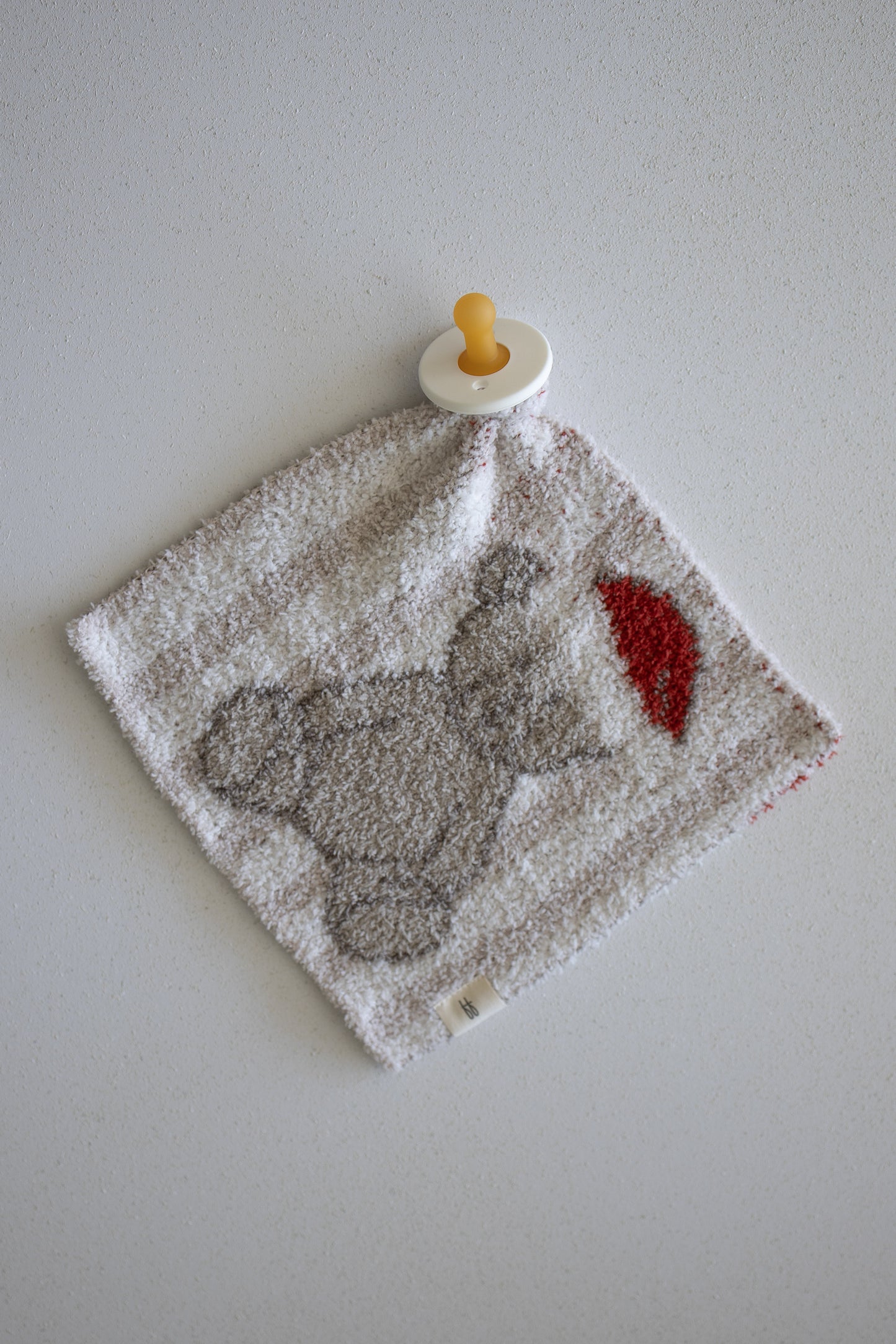 A pacifier is attached to a soft, square baby cloth lovey from forever french baby, called the Lovey | Santa Teddy, featuring a gray elephant holding a red balloon and lying on a light-colored surface.
