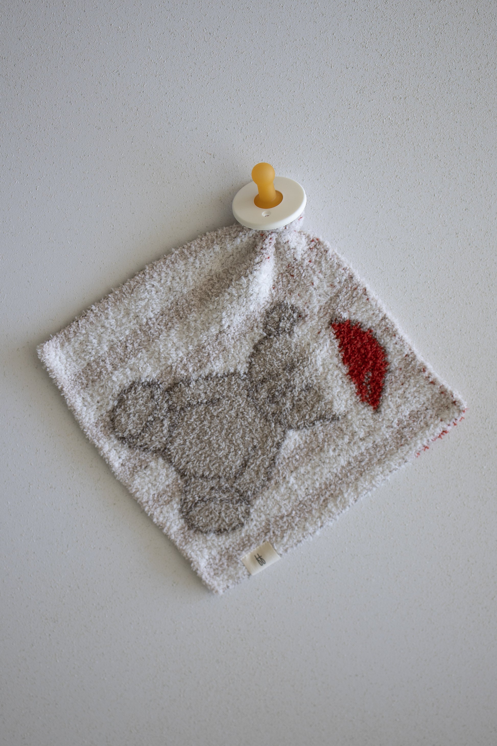 A pacifier is attached to a soft, square baby cloth lovey from forever french baby, called the Lovey | Santa Teddy, featuring a gray elephant holding a red balloon and lying on a light-colored surface.