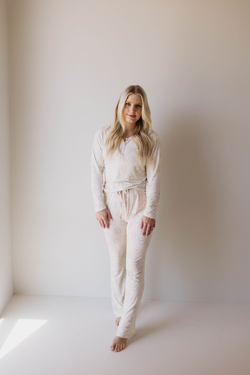 A blonde woman stands barefoot against a plain white background, wearing the "Women's Bamboo Pajamas | Just Smile" from forever french baby—a comfortable white, long-sleeve, button-up pajama set made from bamboo viscose with a subtle pattern. She gazes directly at the camera with a relaxed expression as natural light streams in from the left side.