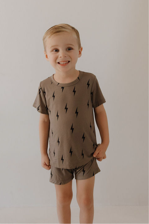 A young child with short blond hair smiles at the camera, wearing the forever french baby Bamboo Short Set in brown with black lightning bolts—soft, hypo-allergenic clothing for sensitive skin—standing against a plain background.