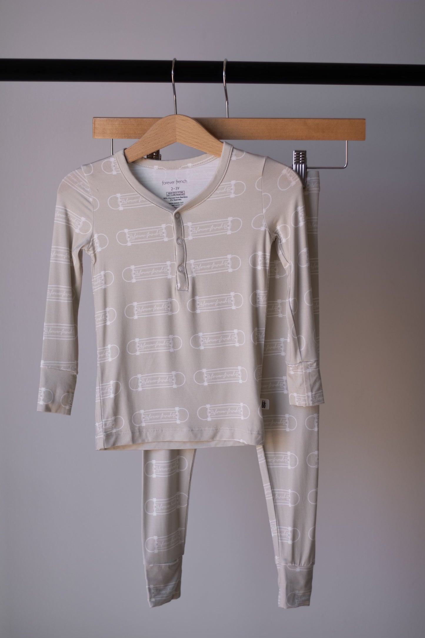 The forever french baby Bamboo Two Piece Pajamas | Sk8er Boy features light beige, breathable bamboo fabric with a subtle skateboard print. This kids’ set includes a long-sleeve top and matching pants, shown hanging on a wooden hanger against a gray wall.