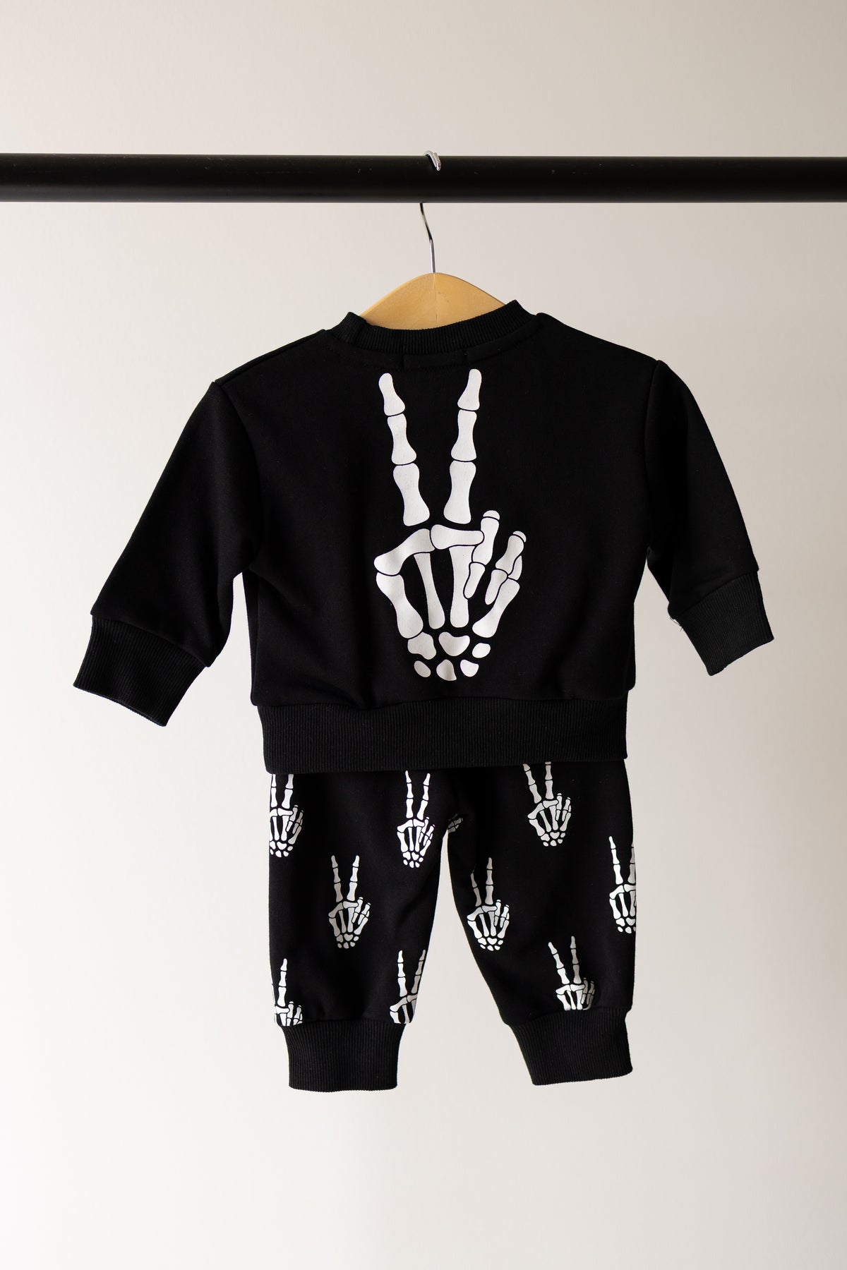 The forever french baby Child Sweat Set | Midnight Skelly is displayed on a hanger against a plain background, featuring a black sweatshirt and pants with skeleton hand peace sign graphics on the back of the top and repeated on the pants—perfect for everyday adventures.
