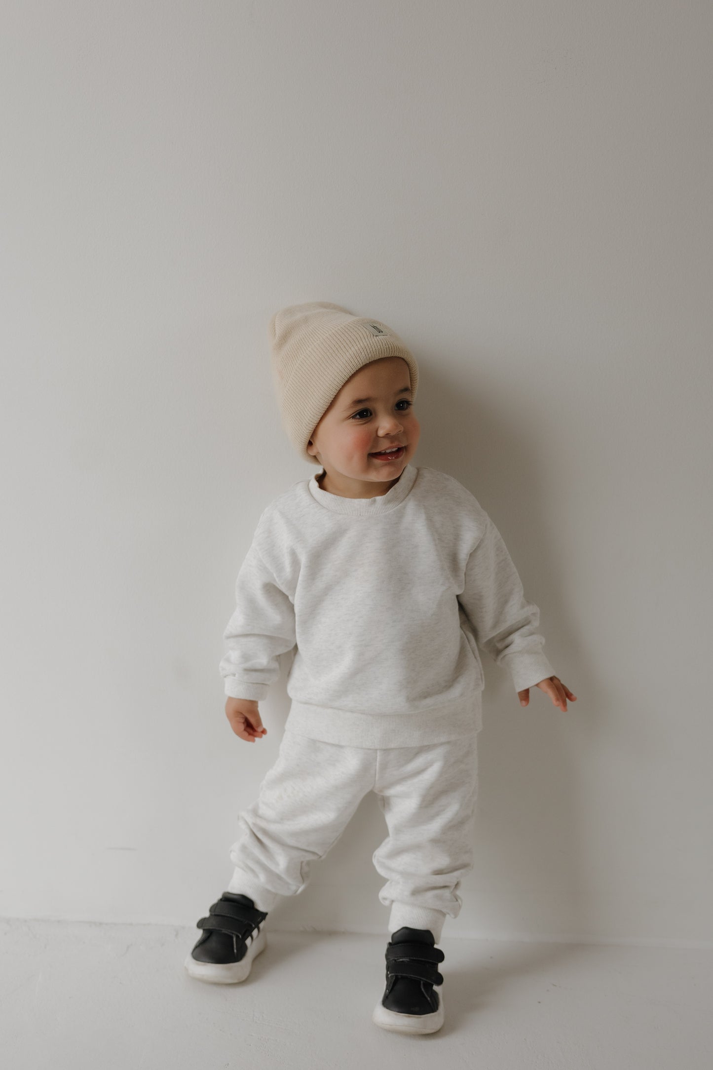 A toddler in the forever french baby Child Sweat Set | Heather Grey—a cozy beanie, sweatshirt, and pants—stands smiling against a white wall. Ideal for comfy days or matching family outfits.