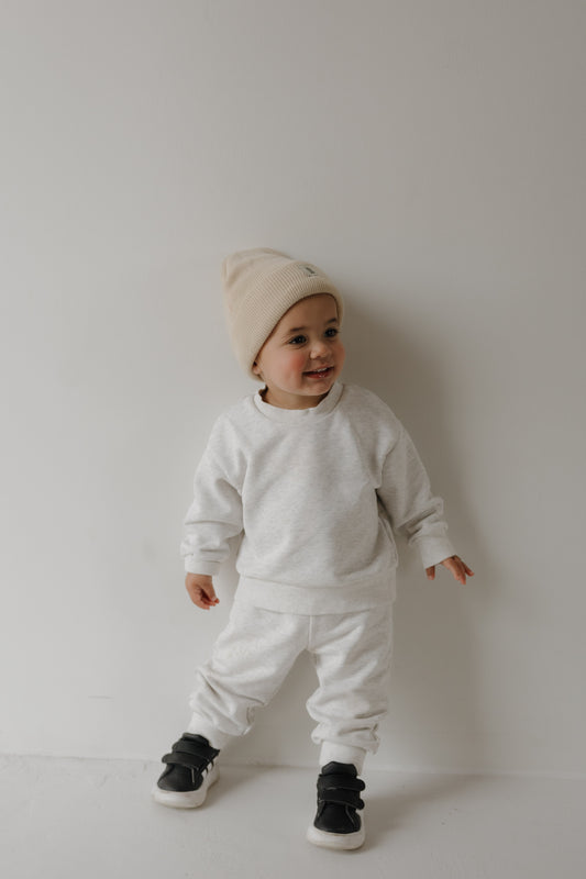 A toddler in the forever french baby Child Sweat Set | Heather Grey—a cozy beanie, sweatshirt, and pants—stands smiling against a white wall. Ideal for comfy days or matching family outfits.