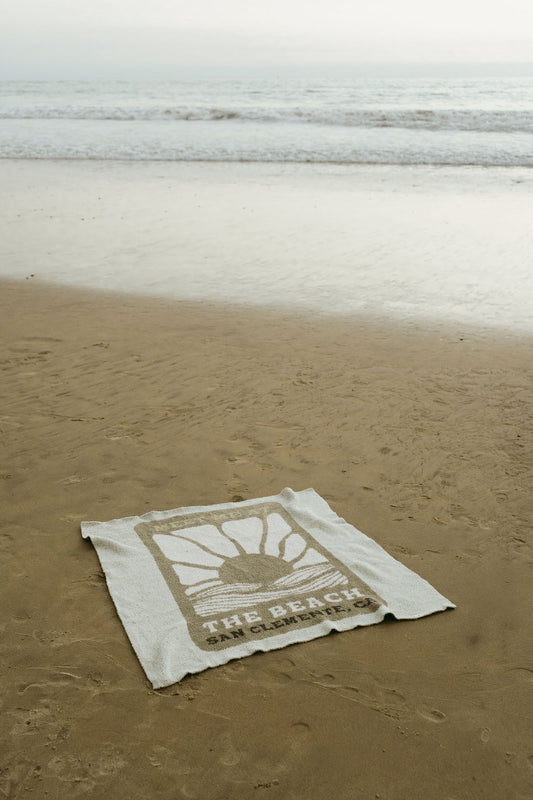 A white OEKO-TEX certified Plush Blanket by forever french baby, featuring a sun and wave design with "Meet Me at the Beach," rests on wet sand near gentle ocean waves beneath an overcast sky.