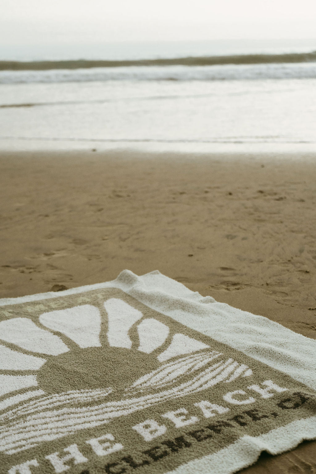 A close-up of the forever french baby Plush Blanket | Meet Me at the Beach, featuring a sun and wave design with "THE BEACH" text, lies on sandy shore near the ocean under a clear sky. OEKO-TEX certified microfiber ensures comfort and quality.