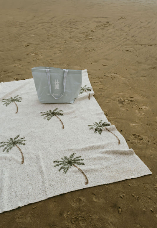 A pale blue beach bag rests on the sand beside a white towel with green palm trees and a cozy forever french baby Plush Blanket | Palm Tree, Oeko-Tex certified. The mostly smooth sand has a few footprints and marks.