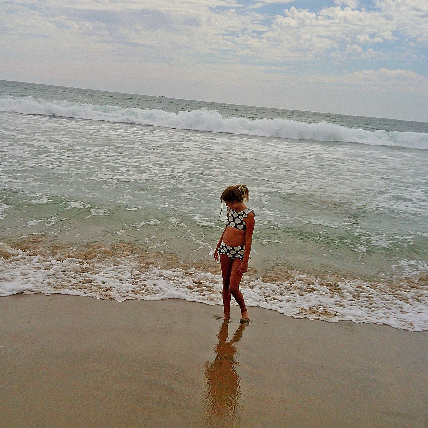 Wearing the forever french baby Girl's Two Piece Swimsuit | Seashells, a girl stands on the sandy beach at the ocean's edge, gazing down as gentle waves approach her feet beneath a partly cloudy sky.