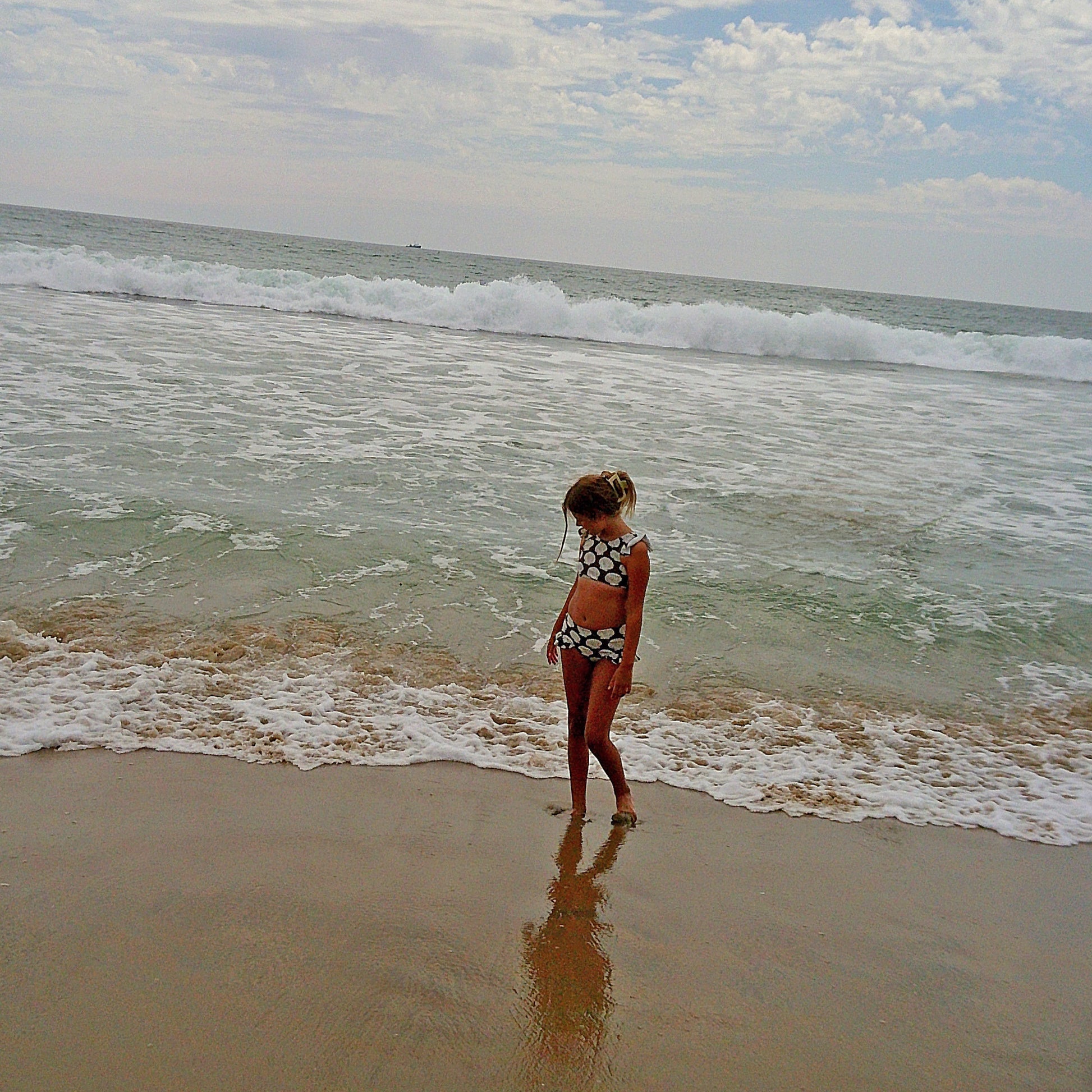 Wearing the forever french baby Girl's Two Piece Swimsuit | Seashells, a girl stands on the sandy beach at the ocean's edge, gazing down as gentle waves approach her feet beneath a partly cloudy sky.