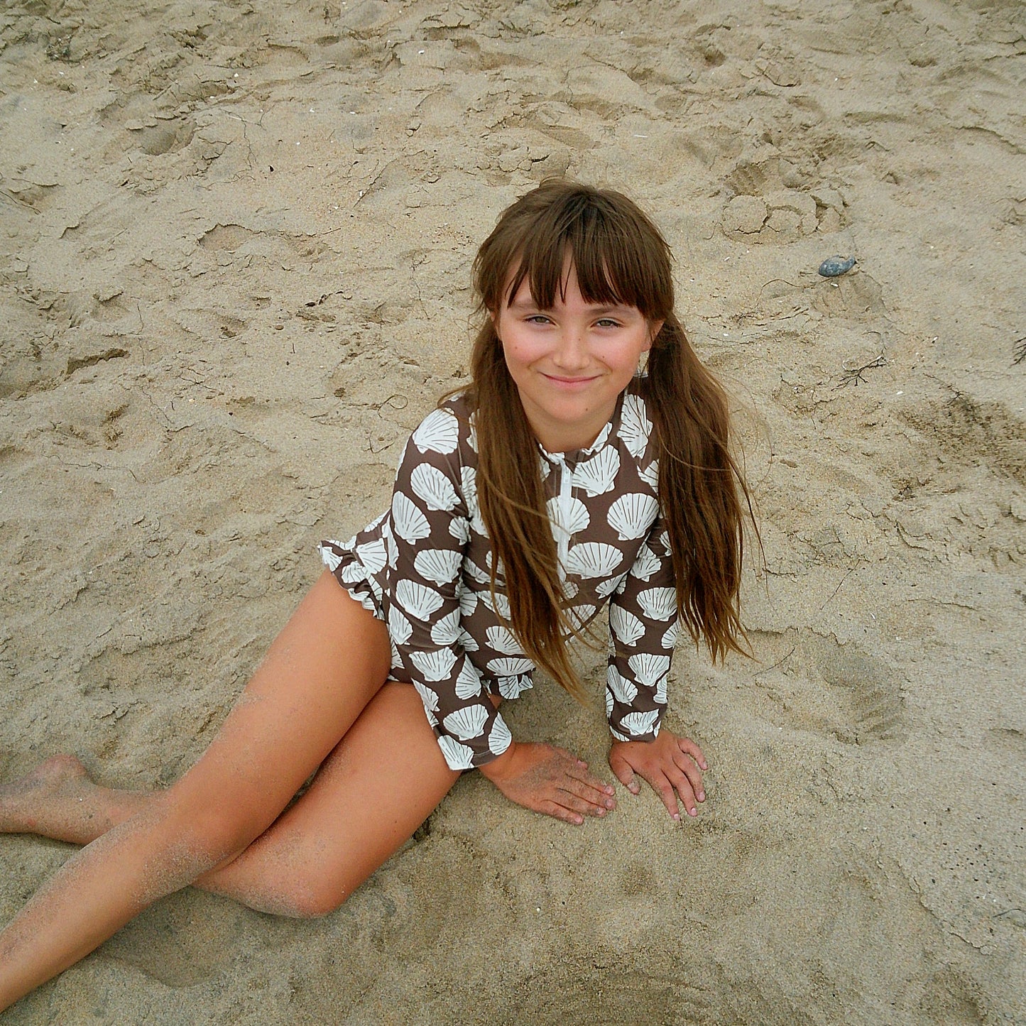 A young girl with long brown hair smiles on the beach, wearing the forever french baby Girl's Long Sleeve Swimsuit | Seashells—a stylish kids' swimwear option with a chic shell print.