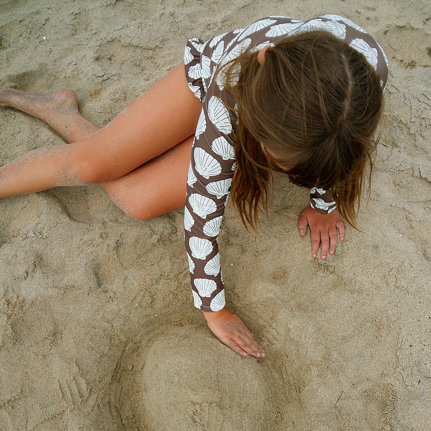 A child with long hair, wearing the forever french baby Girl's Long Sleeve Swimsuit | Seashells, sits on sandy ground shaping the sand with both hands.