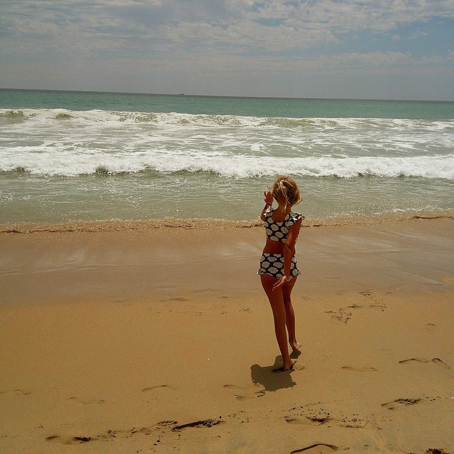 A girl wearing the forever french baby Girl's Two Piece Swimsuit | Seashells stands on a sandy beach near the shoreline, gazing at the ocean waves under a partly cloudy sky.