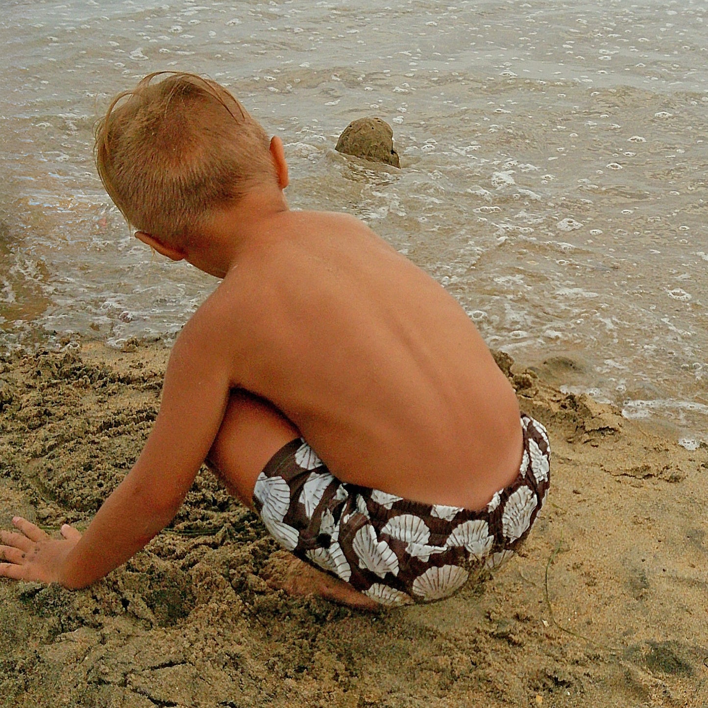 A young child in forever french baby’s Child Board Short | Seashells plays on the sandy beach, crouched near gentle waves and building a small sand mound.