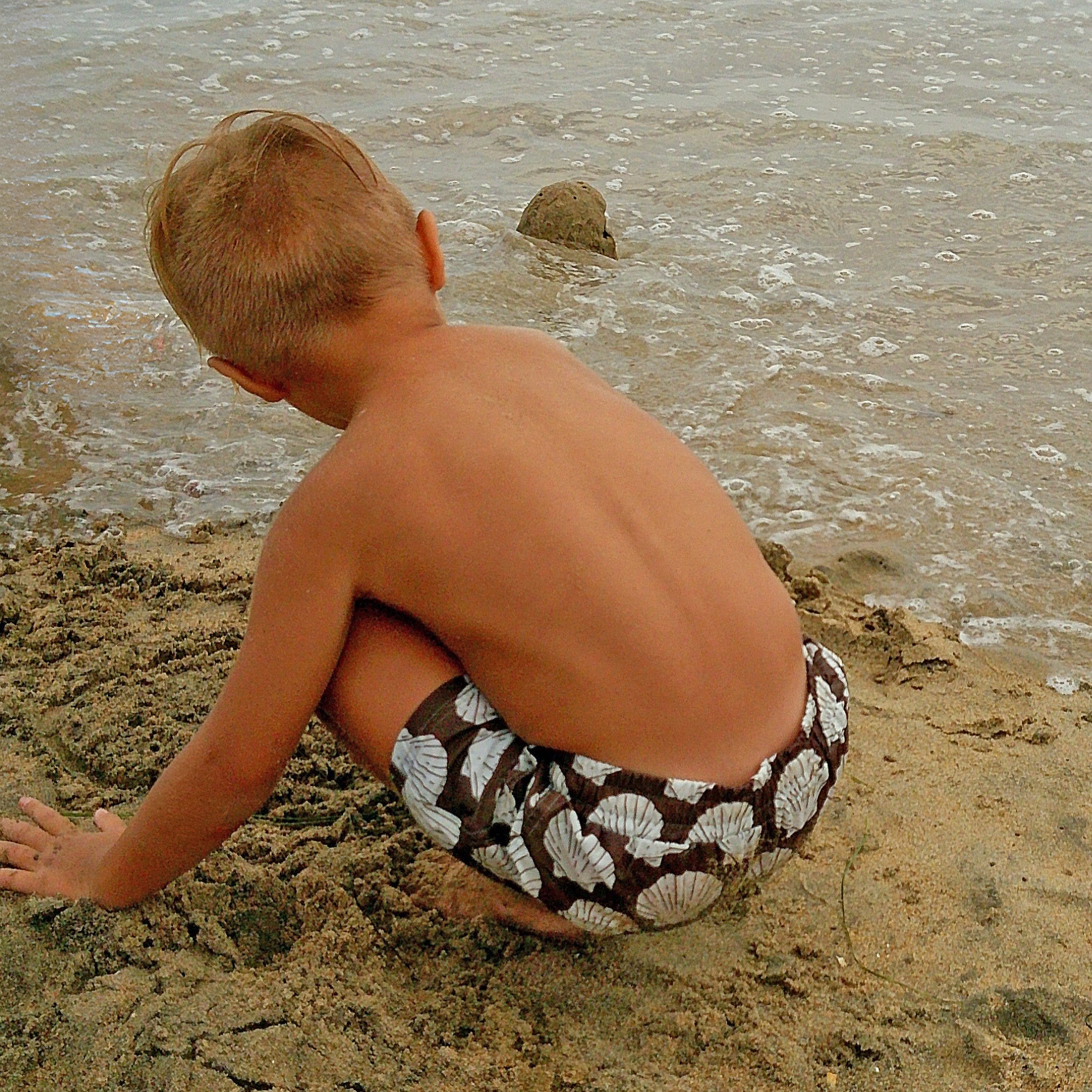 A young child in forever french baby’s Child Board Short | Seashells plays on the sandy beach, crouched near gentle waves and building a small sand mound.