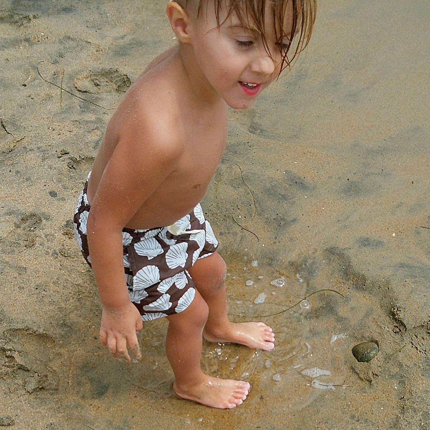 A young child with wet hair stands barefoot on the beach, wearing forever french baby's Child Board Short | Seashells—perfect for summer days and kids’ swimwear moments.
