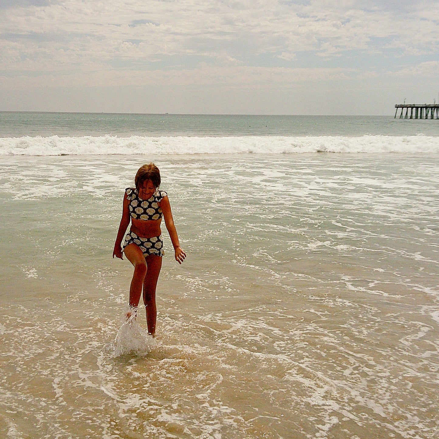 A girl wearing the forever french baby Girl's Two Piece Swimsuit | Seashells walks through shallow waves on a cloudy beach day, with a pier in the background and gentle surf rolling in.