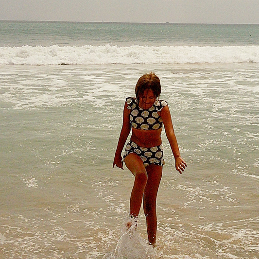 A girl wearing the forever french baby Girl’s Two Piece Swimsuit in Seashells walks through shallow ocean water at the beach, with waves and a cloudy sky in the background.