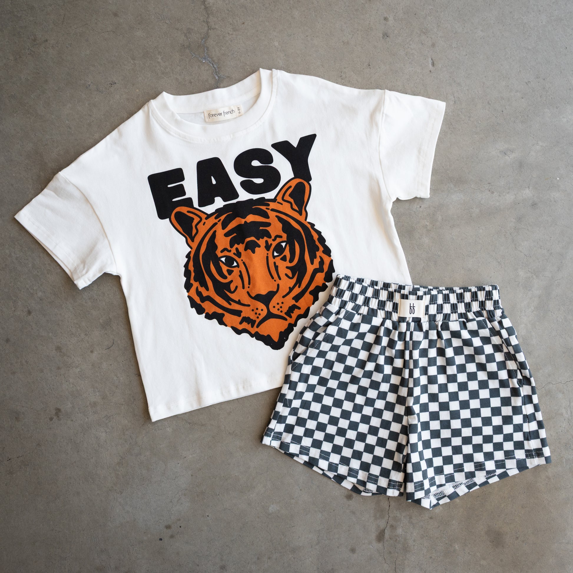 The forever french baby Child Short Set | Easy Tiger features a white cotton-blend tee with a bold tiger face and "EASY" above, paired with black-and-white checkered shorts, displayed on a gray concrete surface.