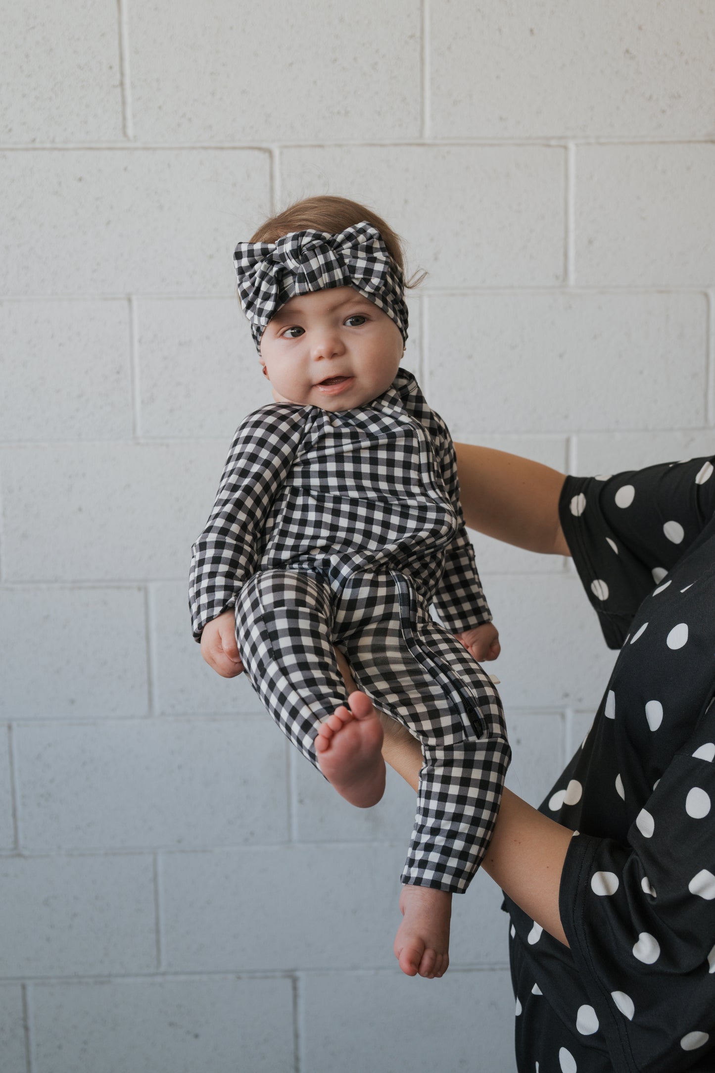 An adult dressed in black with white polka dots holds a baby wearing the forever french baby Bamboo Head Wrap in Black Gingham, set against a light brick wall background.