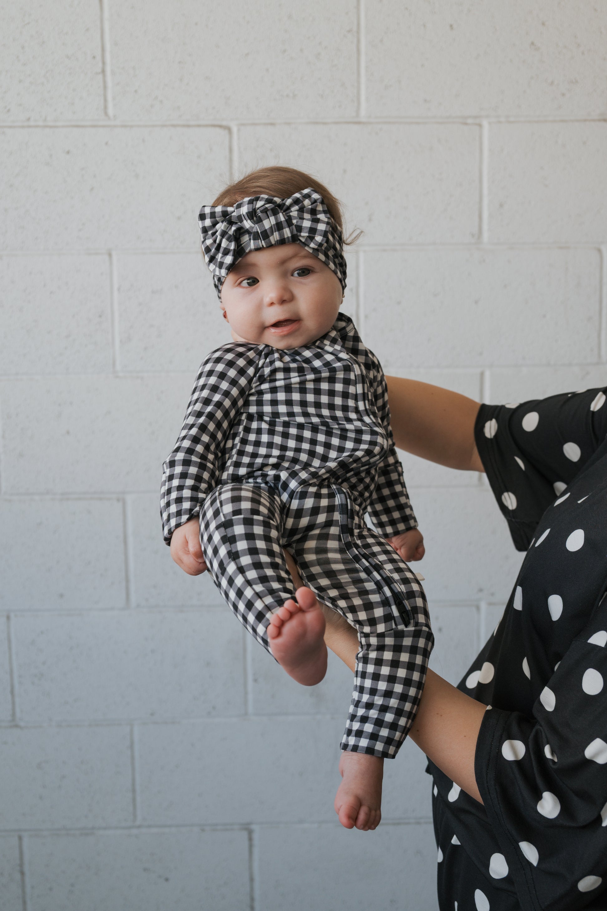 An adult dressed in black with white polka dots holds a baby wearing the forever french baby Bamboo Head Wrap in Black Gingham, set against a light brick wall background.