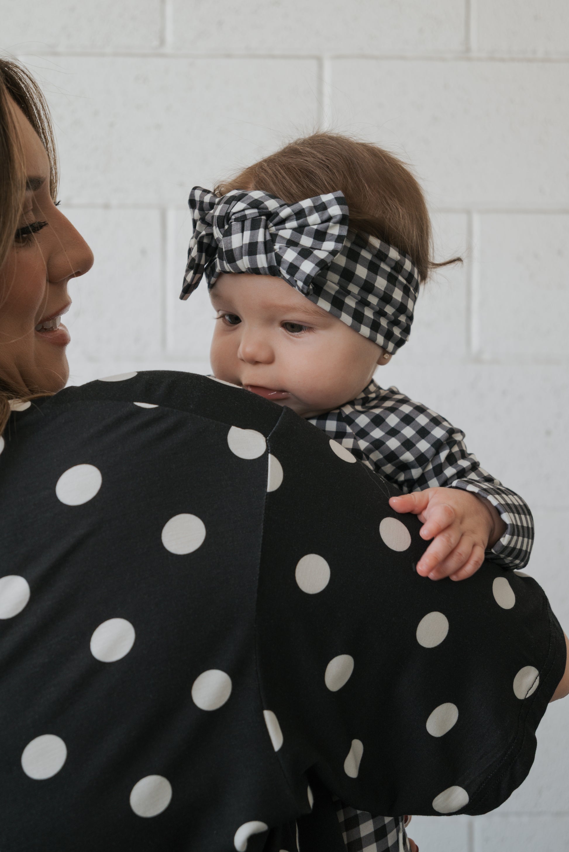 A baby rests on a woman’s shoulder, wearing the forever french baby Bamboo Head Wrap in Black Gingham and a matching outfit. The woman is dressed in a black top with large white polka dots. They are indoors, set against a white brick wall.