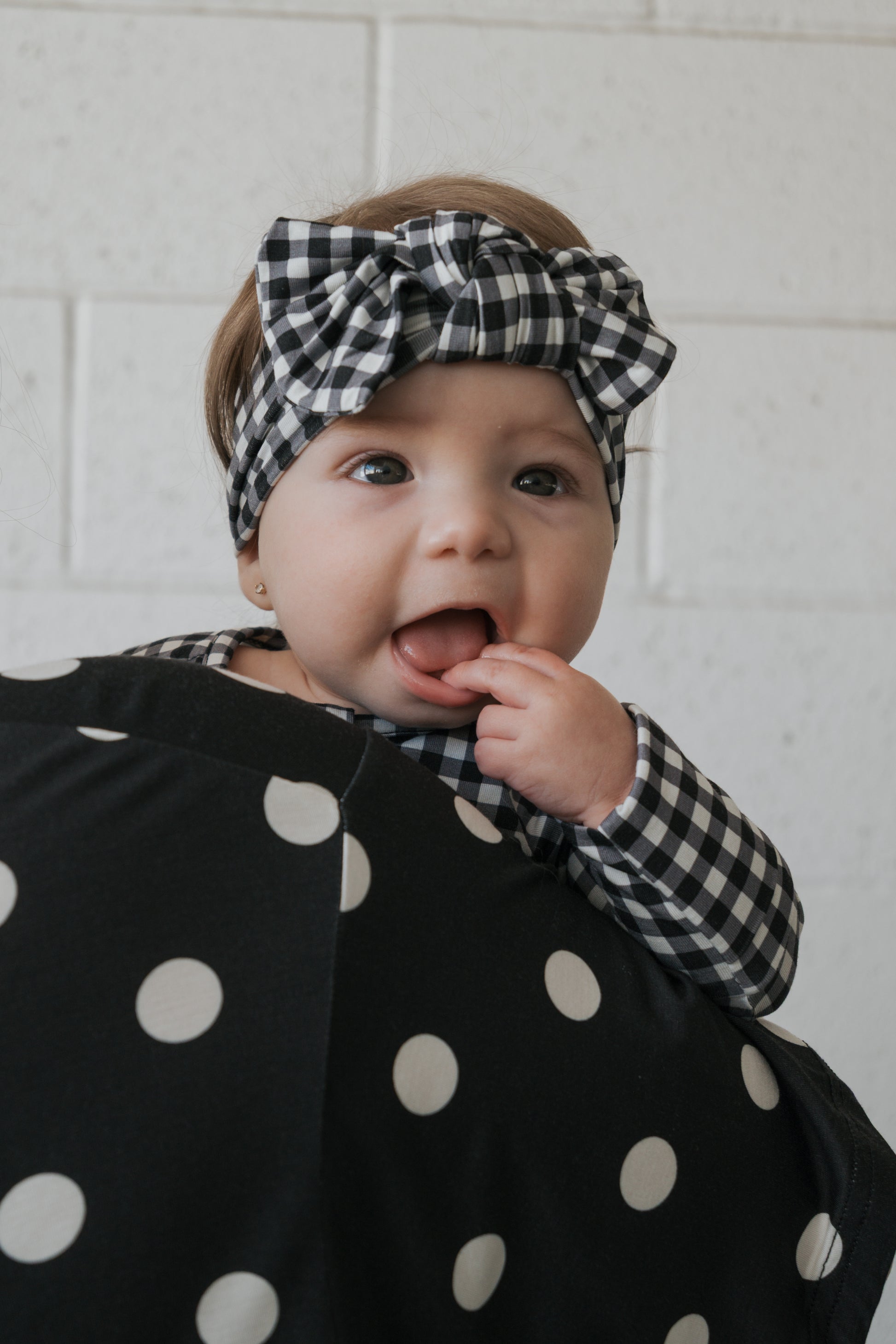 Wearing the Bamboo Head Wrap in Black Gingham by forever french baby, a baby dressed in a matching outfit touches their mouth while leaning against a person draped in black-and-white polka dot fabric.