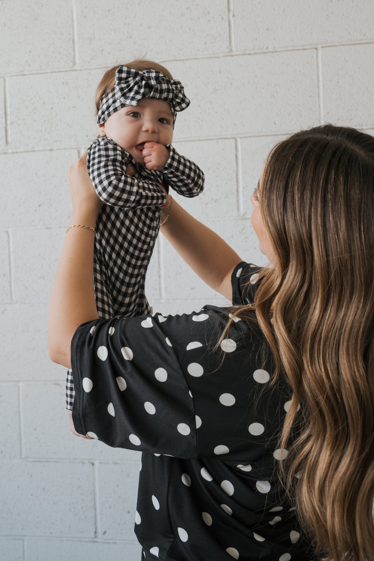 A woman with long wavy hair holds a baby wearing the forever french baby Bamboo Head Wrap in Black Gingham, along with a matching gingham outfit, against a white brick wall. The woman is dressed in a black top with white polka dots.