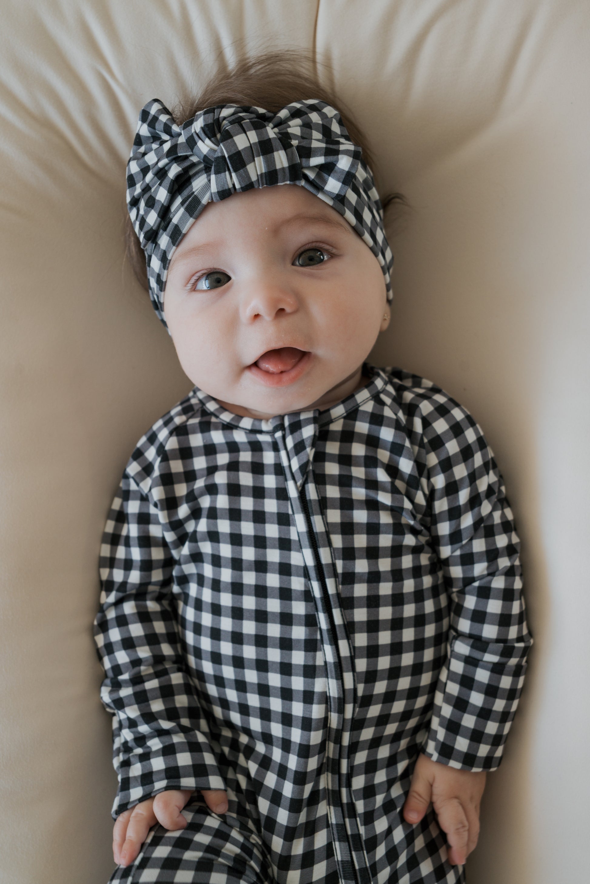 A baby wearing the forever french baby Bamboo Head Wrap in Black Gingham, along with a matching checkered outfit, lies on a light-colored surface, gazing upward with their tongue slightly out.