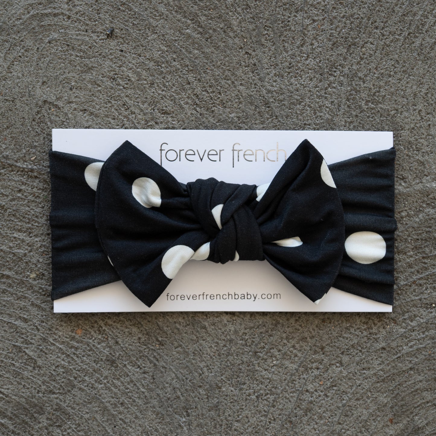 Showcased on a textured gray surface, the Bamboo Head Wrap | B&W Polka Dot from forever french baby features bold white polka dots on black and is styled with an adorable bow, presented on a branded “forever french” card.