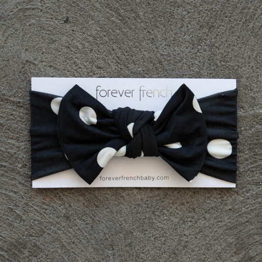Showcased on a textured gray surface, the Bamboo Head Wrap | B&W Polka Dot from forever french baby features bold white polka dots on black and is styled with an adorable bow, presented on a branded “forever french” card.