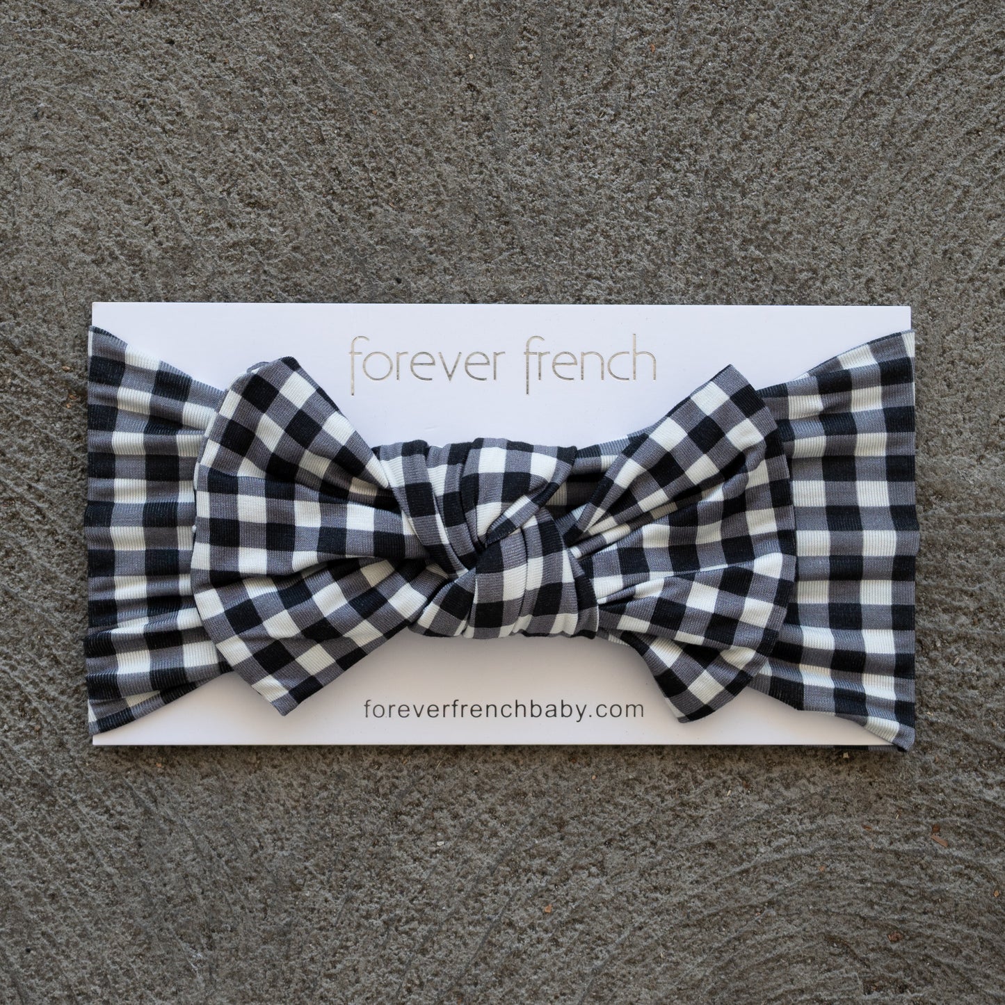 A Bamboo Head Wrap in Black Gingham from forever french baby is shown with a bow, displayed on a branded card atop a textured gray surface.