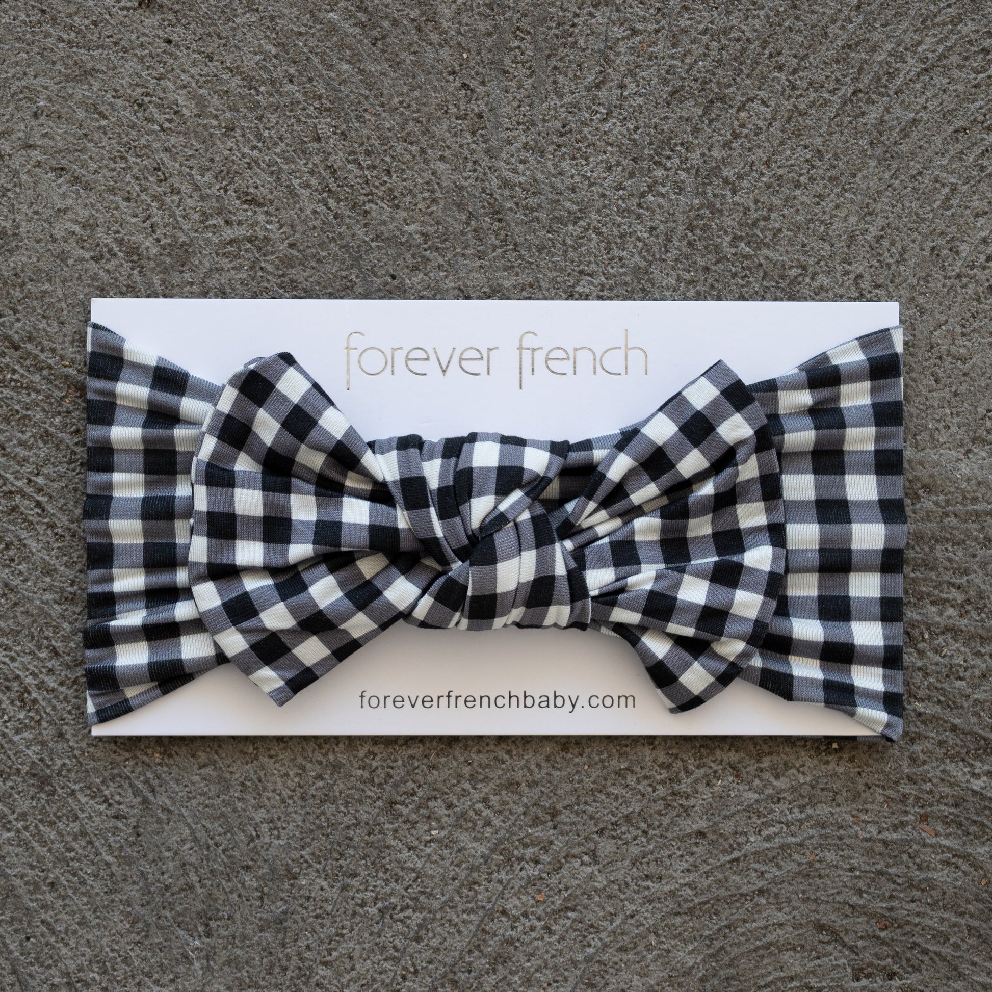 A Bamboo Head Wrap in Black Gingham from forever french baby is shown with a bow, displayed on a branded card atop a textured gray surface.