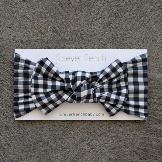 A Bamboo Head Wrap in Black Gingham from forever french baby is shown with a bow, displayed on a branded card atop a textured gray surface.