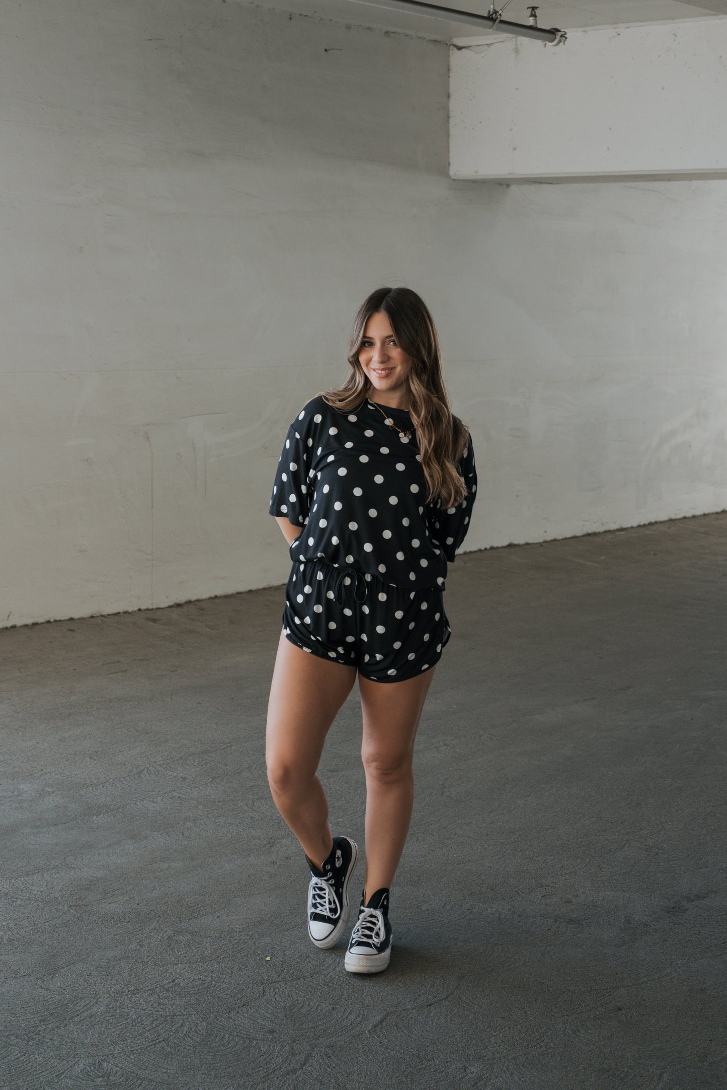 A woman stands indoors on a concrete floor, smiling with her hands behind her back, wearing the forever french baby Women’s Bamboo Short Set in B&W Polka Dot—crafted from breathable bamboo fabric—and black sneakers. The minimal setting features plain white walls.