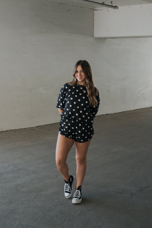 A woman stands indoors on a concrete floor, smiling with her hands behind her back, wearing the forever french baby Women’s Bamboo Short Set in B&W Polka Dot—crafted from breathable bamboo fabric—and black sneakers. The minimal setting features plain white walls.