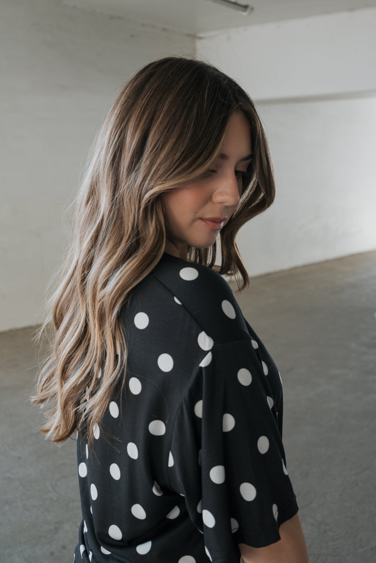 A woman with long, wavy hair wears the forever french baby Women’s Bamboo Short Set in Black & White Polka Dot, looking down in a minimalist, empty room with white walls.